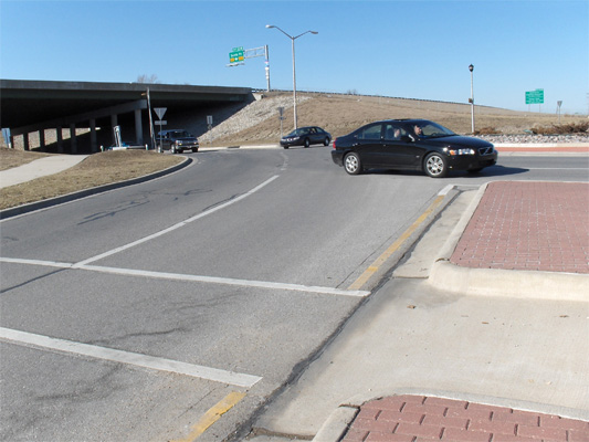 In these two photos, a crosswalk crosses two lanes from an island in the middle of the street to the sidewalk.  About 20 feet to our right, our street enters a roundabout (lanes go around a large circle) with another street entering the roundabout from under a bridge.  There is a very gentle curve such that drivers coming from under the bridge do not need to slow down much to make the turn into our street.  The two photos show cars coming from under the bridge and from the other side of the circle, with one car going past our street to continue around the circle.